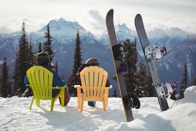 Free photo couple sitting on chair by snowboards at snow covered mountain